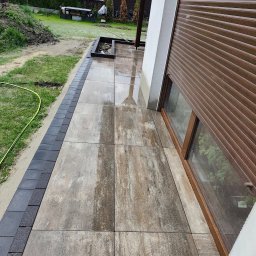 BRUKIO MICHAŁ JABŁOŃSKI - Ułożony taras z płytek przy domu z roletą zewnętrzną. Widoczne obrzeża z kostki. Częściowo mokra nawierzchnia. Prace wykończeniowe w ogrodzie w tle.