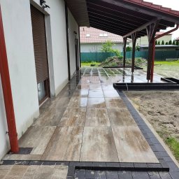 BRUKIO MICHAŁ JABŁOŃSKI - Wyłożony płytami taras przy domu z zadaszeniem, obrzeża z ciemnej kostki. Widoczne prace ziemne w ogrodzie. Po deszczu, mokra nawierzchnia.