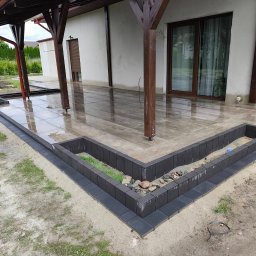 BRUKIO MICHAŁ JABŁOŃSKI - Wykończony taras z płytek, obrzeża z kostki w formie rabaty z kamieniami i zielenią. Zadaszenie z drewnianymi słupami. Widok na dom i ogród w tle.