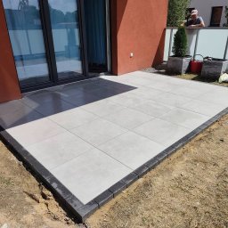 BRUKIO MICHAŁ JABŁOŃSKI - Wykończony taras z jasnych płyt, obramowany ciemną kostką, przylegający do budynku z dużymi oknami. W tle widoczny fragment ogrodzenia i osoba.