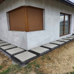 BRUKIO MICHAŁ JABŁOŃSKI - Nowoczesna ścieżka z kwadratowych płyt betonowych ułożona wzdłuż surowej ściany domu z brązowymi roletami okiennymi, otoczona ziemią z trawą.