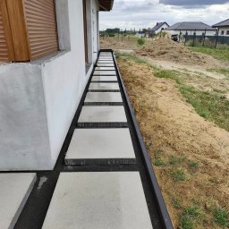 BRUKIO MICHAŁ JABŁOŃSKI - Nowoczesna ścieżka z kwadratowych płyt betonowych, ułożonych wzdłuż budynku z szarą elewacją. Czarny żwir i obrzeża. W tle widok na domy i pochmurne niebo.