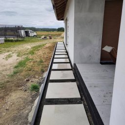 BRUKIO MICHAŁ JABŁOŃSKI - Nowoczesna ścieżka z jasnych płyt betonowych, obrzeża z ciemnej kostki, prowadząca do wejścia domu. W tle trawnik i częściowo widoczny trampolina.