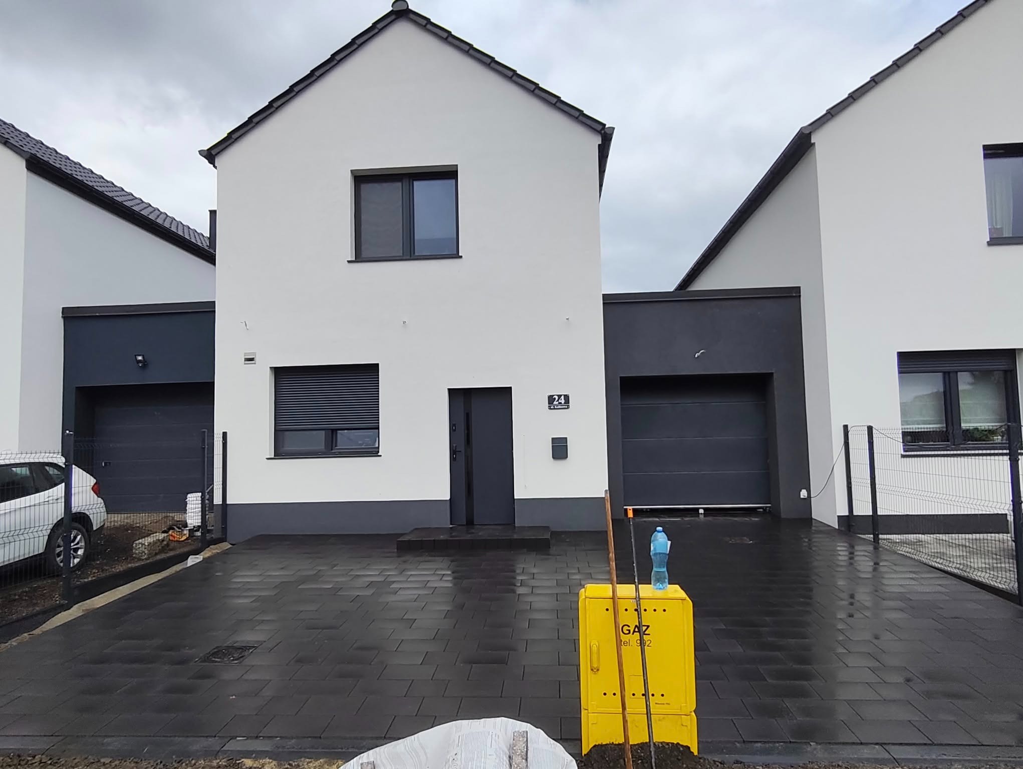 Nowoczesny podjazd z ciemnej kostki brukowej przed domem o białej elewacji i grafitowych akcentach. Żółta skrzynka gazowa na pierwszym planie.