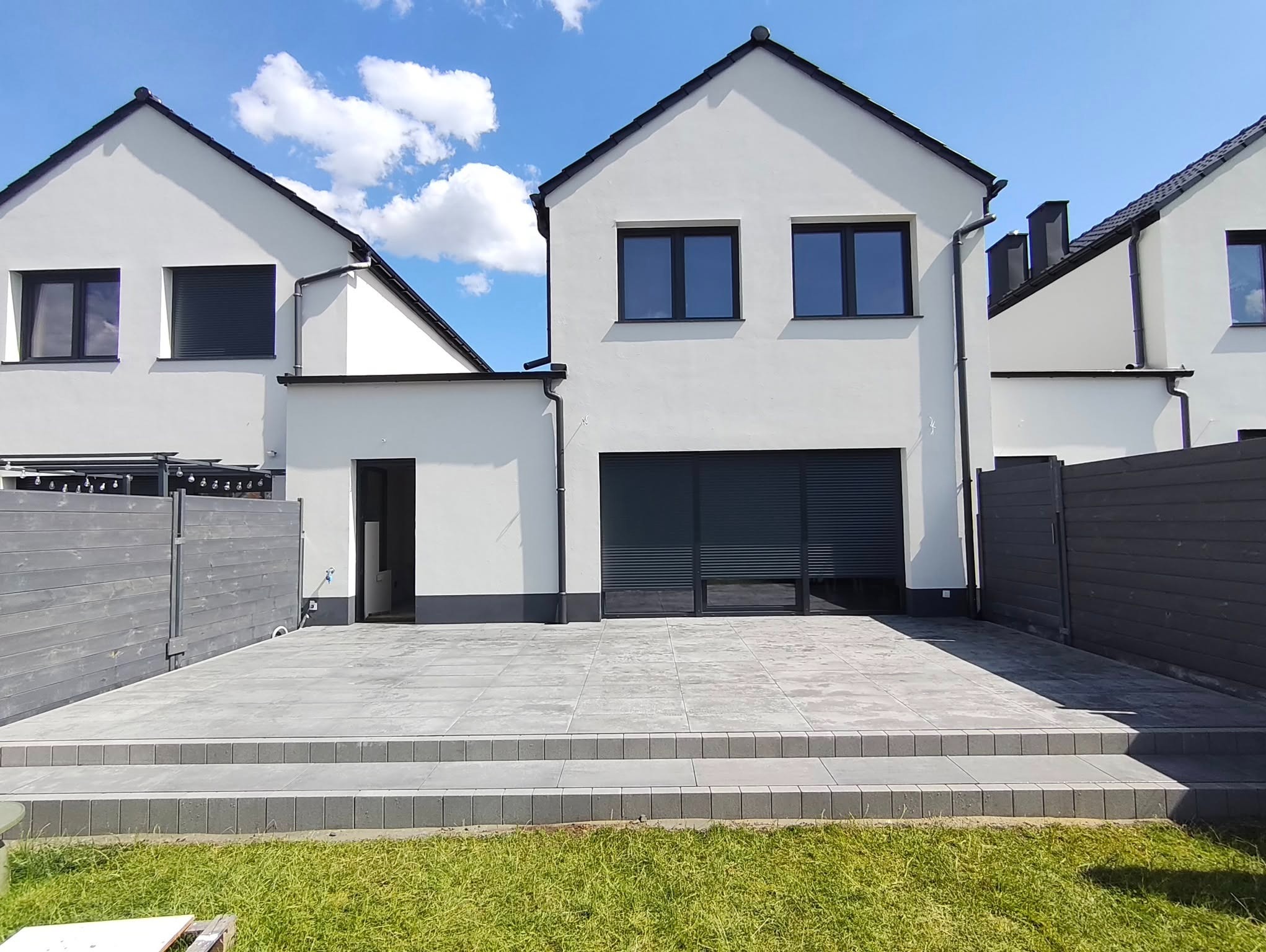 Nowoczesny taras z szarych płyt, schody z kostki, białe domy w tle, błękitne niebo. Minimalistyczny design, świeżo po realizacji, schludne wykończenie.