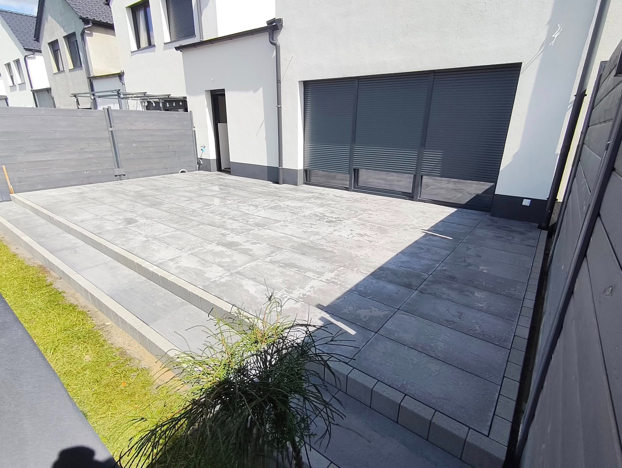 Nowoczesny taras z szarych płyt betonowych, z dwoma stopniami i drewnianym ogrodzeniem w tle, przylegający do białego budynku z roletami.