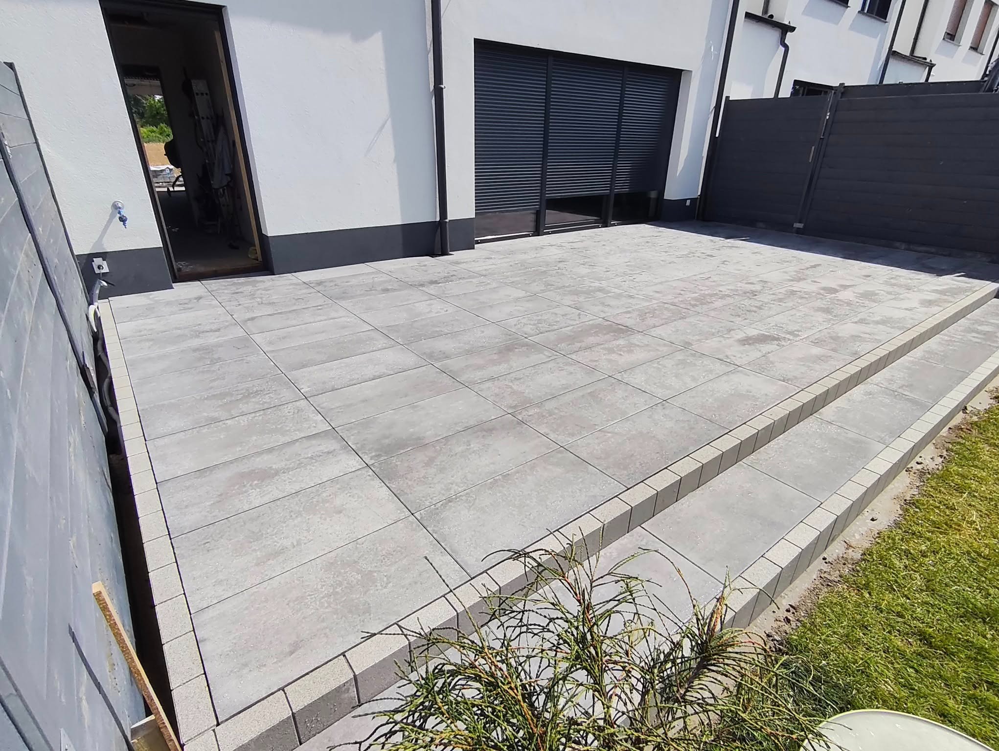 Nowoczesny taras z szarych płyt betonowych, z widocznymi schodkami i fragmentem zieleni. Minimalistyczny styl, jasne oświetlenie, schludne wykończenie.