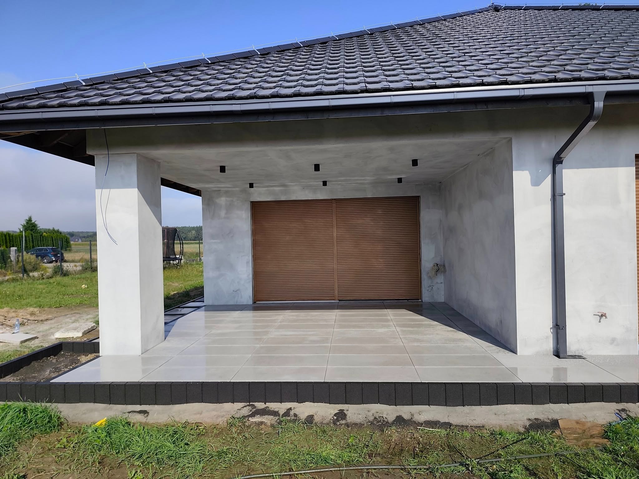 Zadaszony taras z szarych płyt, wykończony obrzeżem z ciemnej kostki. Widoczne kolumny podtrzymujące dach i roleta garażowa. W tle zieleń i błękitne niebo.
