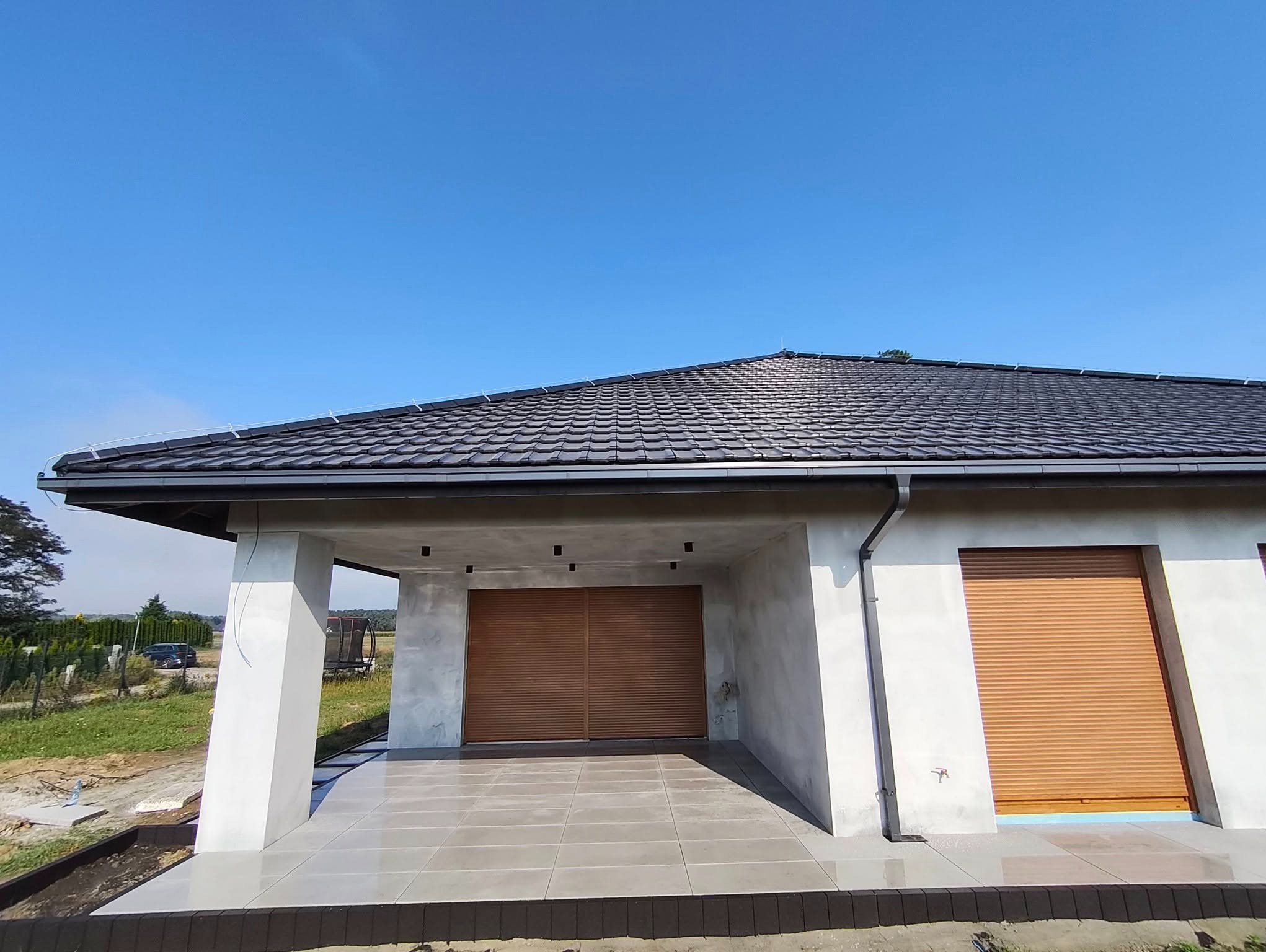 Nowoczesny dom z tarasem wyłożonym szarymi płytkami, brązowe rolety okienne i garażowe, czarny dach, błękitne niebo w tle. Widok z dołu.