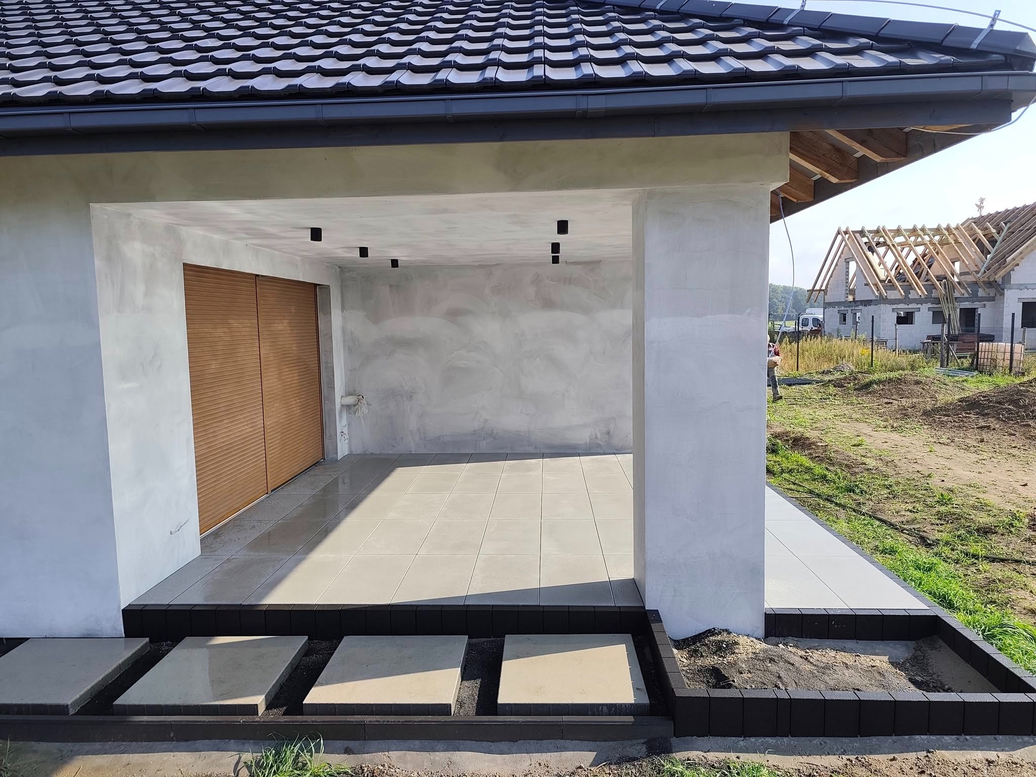 Zadaszone patio z jasnymi płytkami podłogowymi i drewnianymi roletami, obok ścieżka z kamiennych płyt. W tle budowa domu z widoczną więźbą dachową.