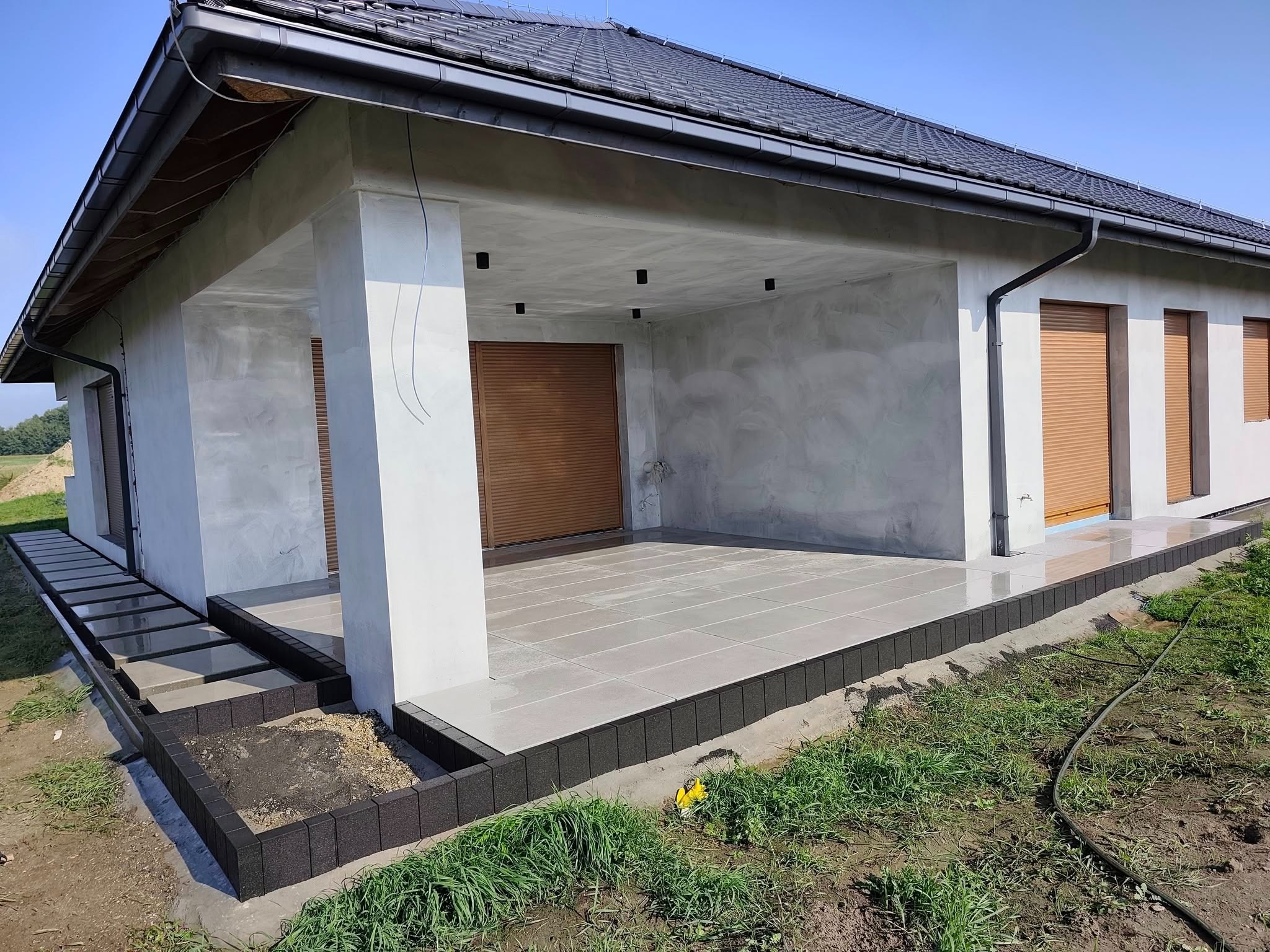 Zadaszony taras z szarych płyt, obrzeża z ciemnej kostki. Widoczne schody wejściowe i fragment trawnika. Szare ściany budynku z roletami.