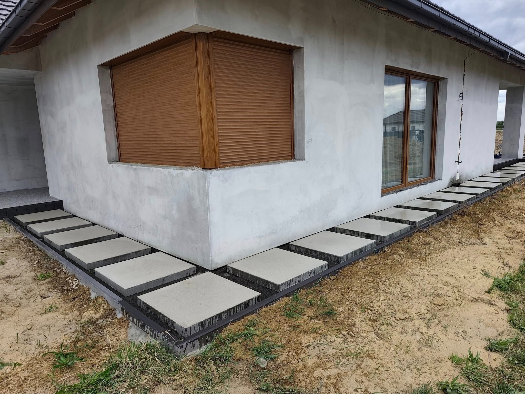 Nowoczesna ścieżka z kwadratowych płyt betonowych ułożona wzdłuż surowej ściany domu z brązowymi roletami okiennymi, otoczona ziemią z trawą.