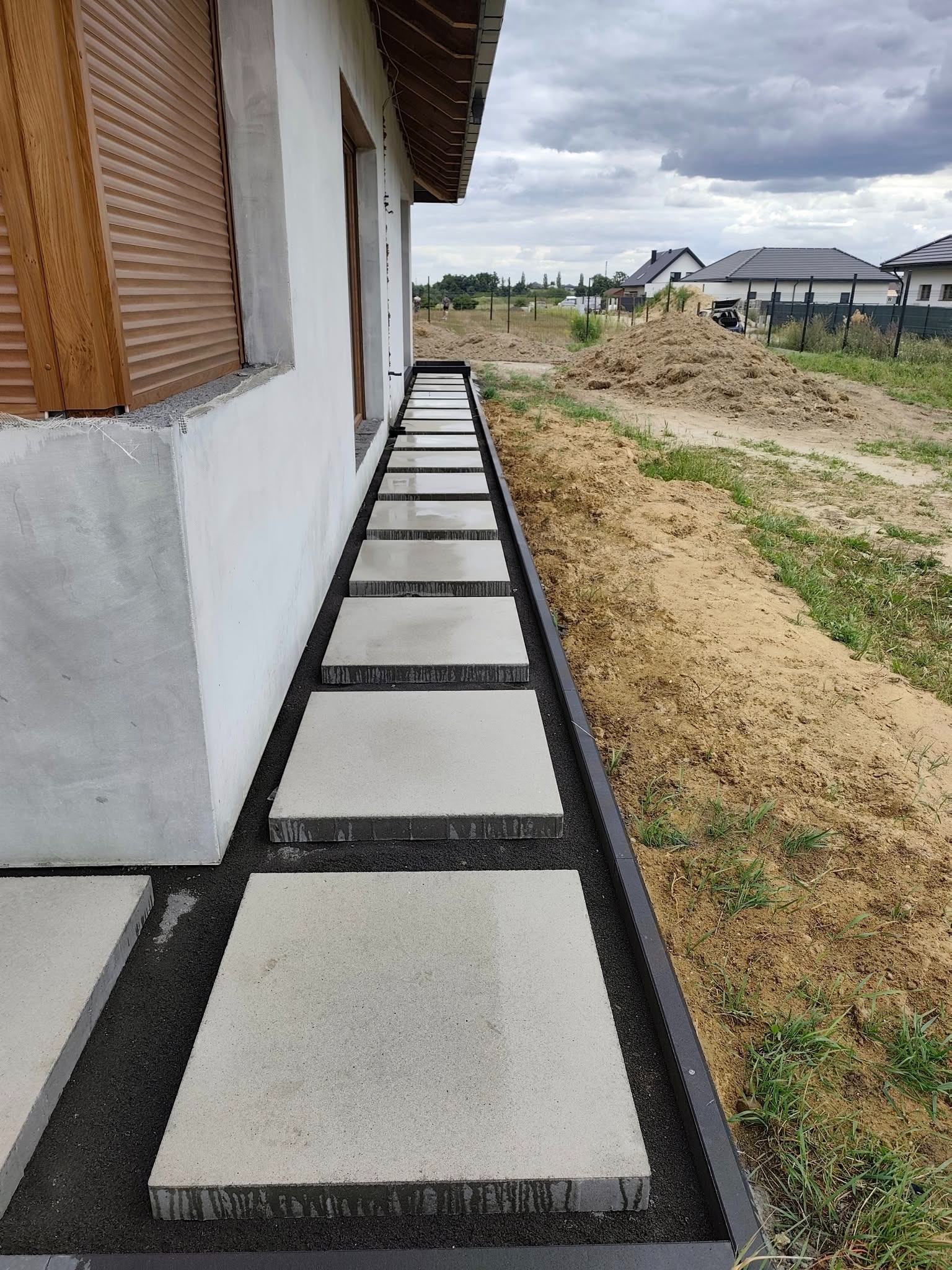 Nowoczesna ścieżka z kwadratowych płyt betonowych, ułożonych wzdłuż budynku z szarą elewacją. Czarny żwir i obrzeża. W tle widok na domy i pochmurne niebo.