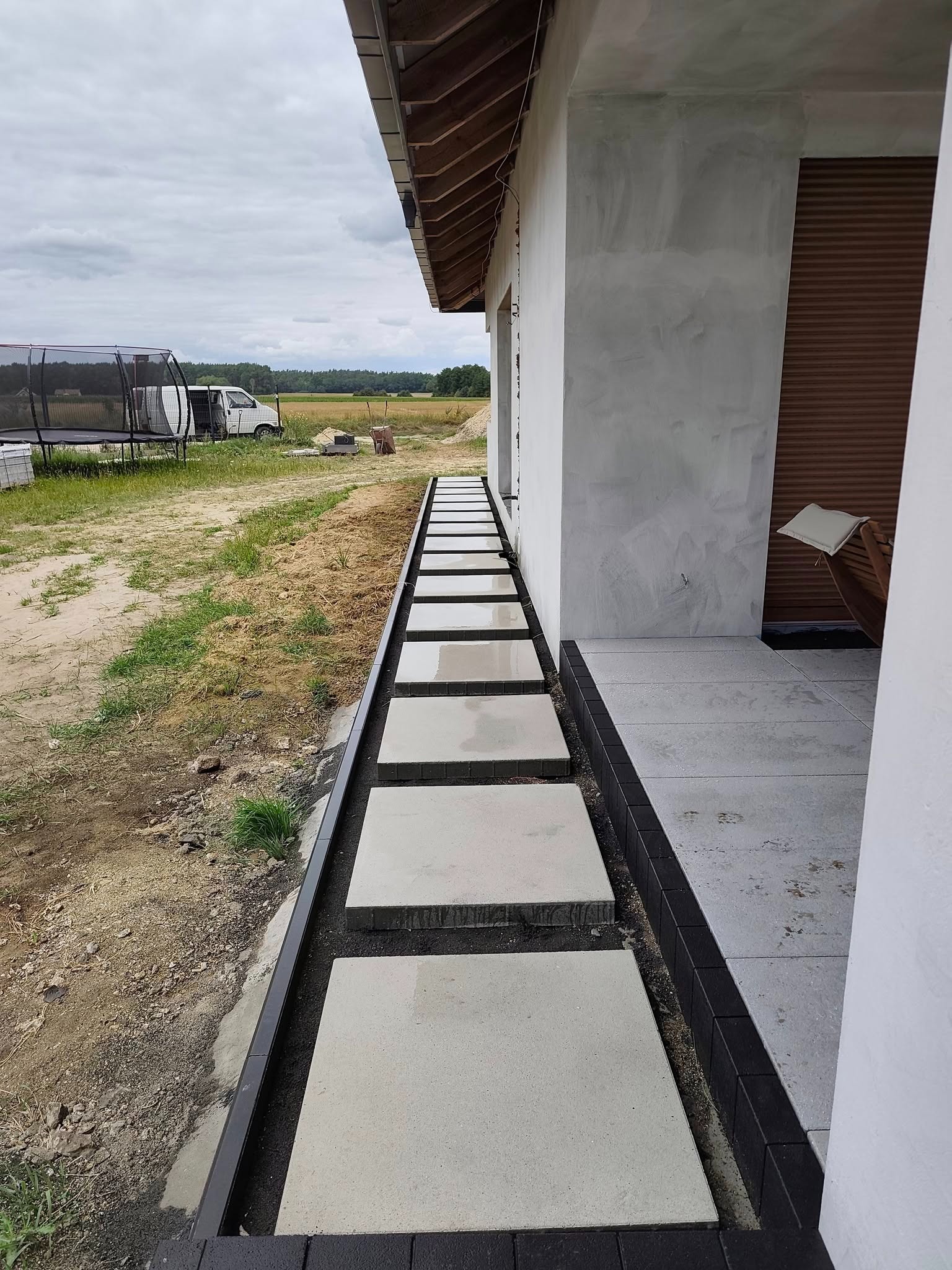 Nowoczesna ścieżka z jasnych płyt betonowych, obrzeża z ciemnej kostki, prowadząca do wejścia domu. W tle trawnik i częściowo widoczny trampolina.