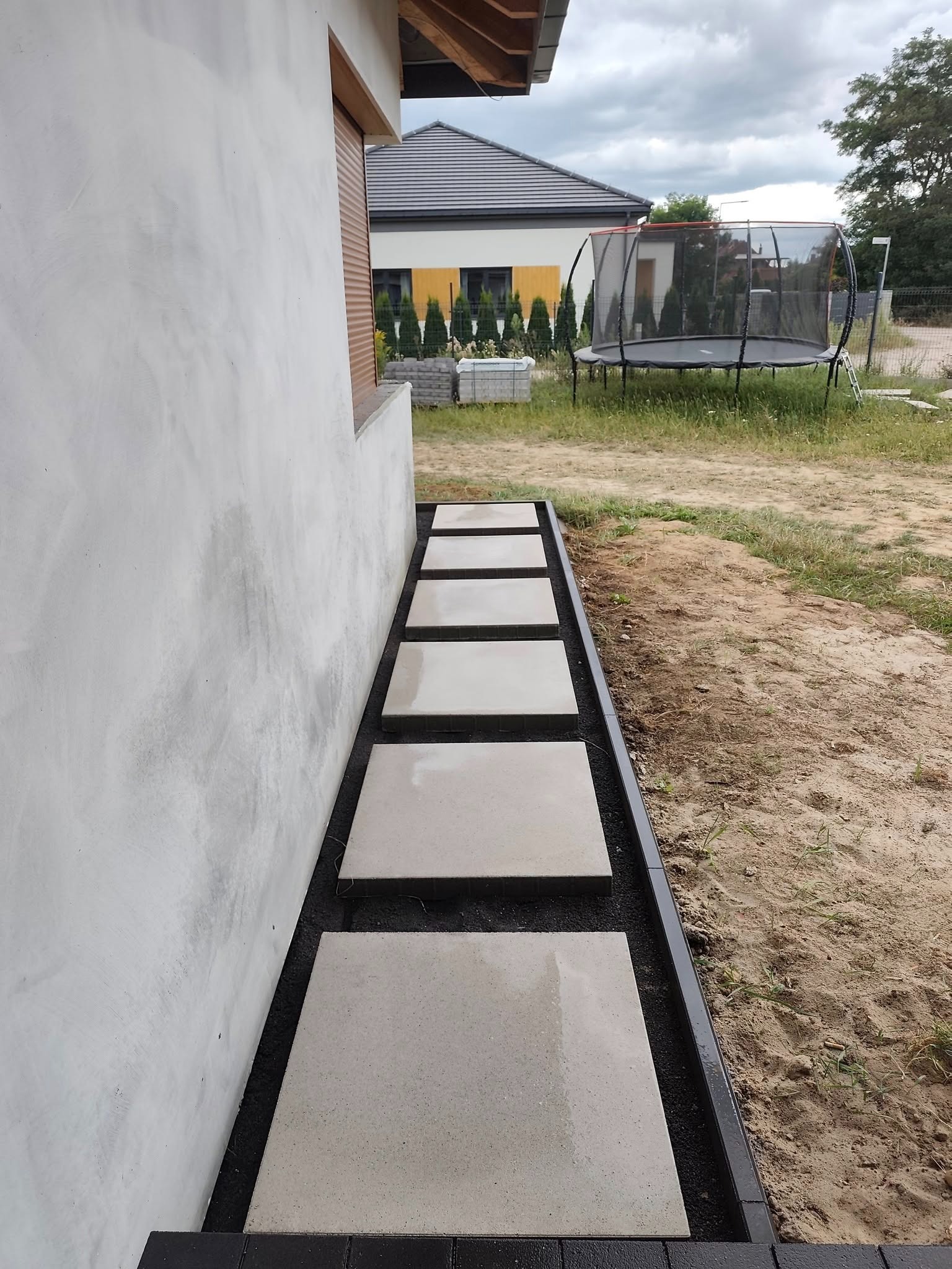 Nowoczesna ścieżka z jasnych płyt betonowych, obramowana czarnym obrzeżem, prowadząca wzdłuż ściany domu. W tle widoczny trawnik i trampolina.