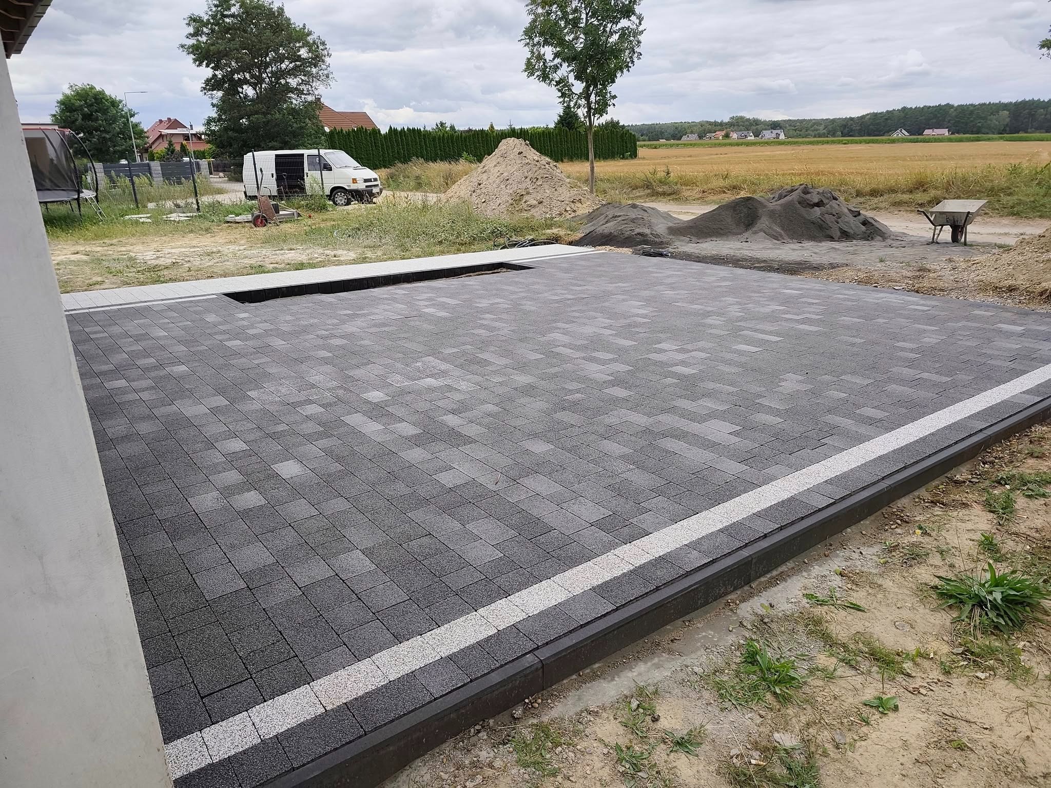 Ułożona szara kostka brukowa z obrzeżem i białym pasem, widok z góry. W tle piasek, gruz i krajobraz wiejski. Częściowy widok ściany budynku po lewej.