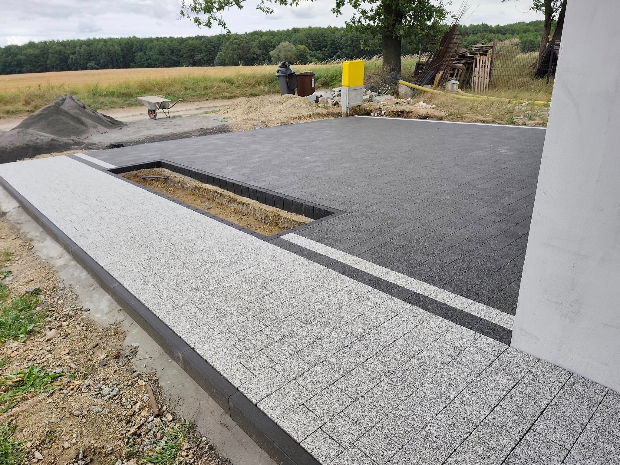 Ułożona kostka brukowa w dwóch odcieniach szarości, z widocznym fragmentem przygotowanego wykopu. W tle widać krajobraz polny z drzewami. Praca w toku.