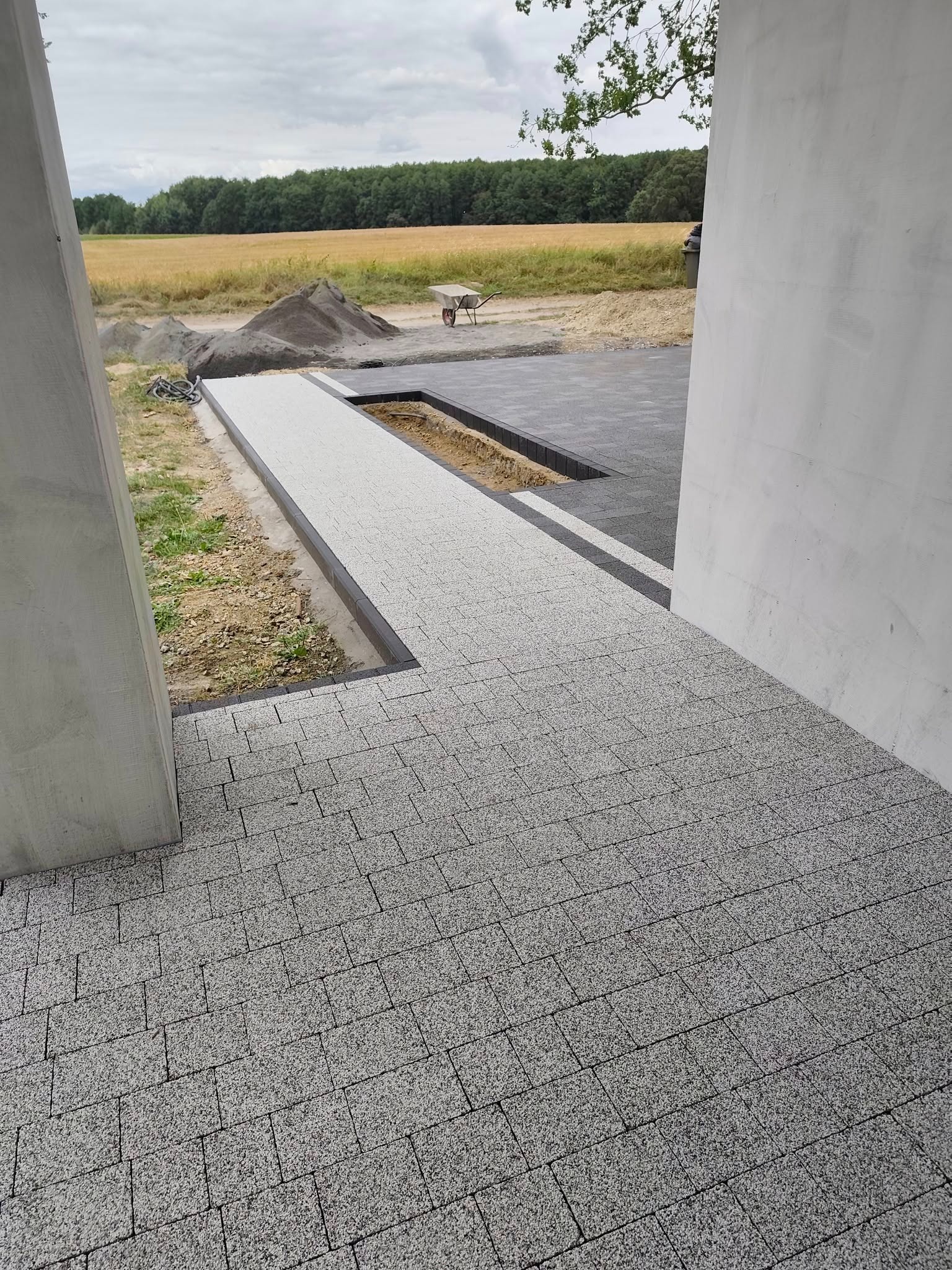 Ułożona kostka brukowa w odcieniach szarości tworząca ścieżkę. W tle widoczne przygotowane materiały i krajobraz polny z drzewami. Kąt ujęcia z perspektywy pieszego.