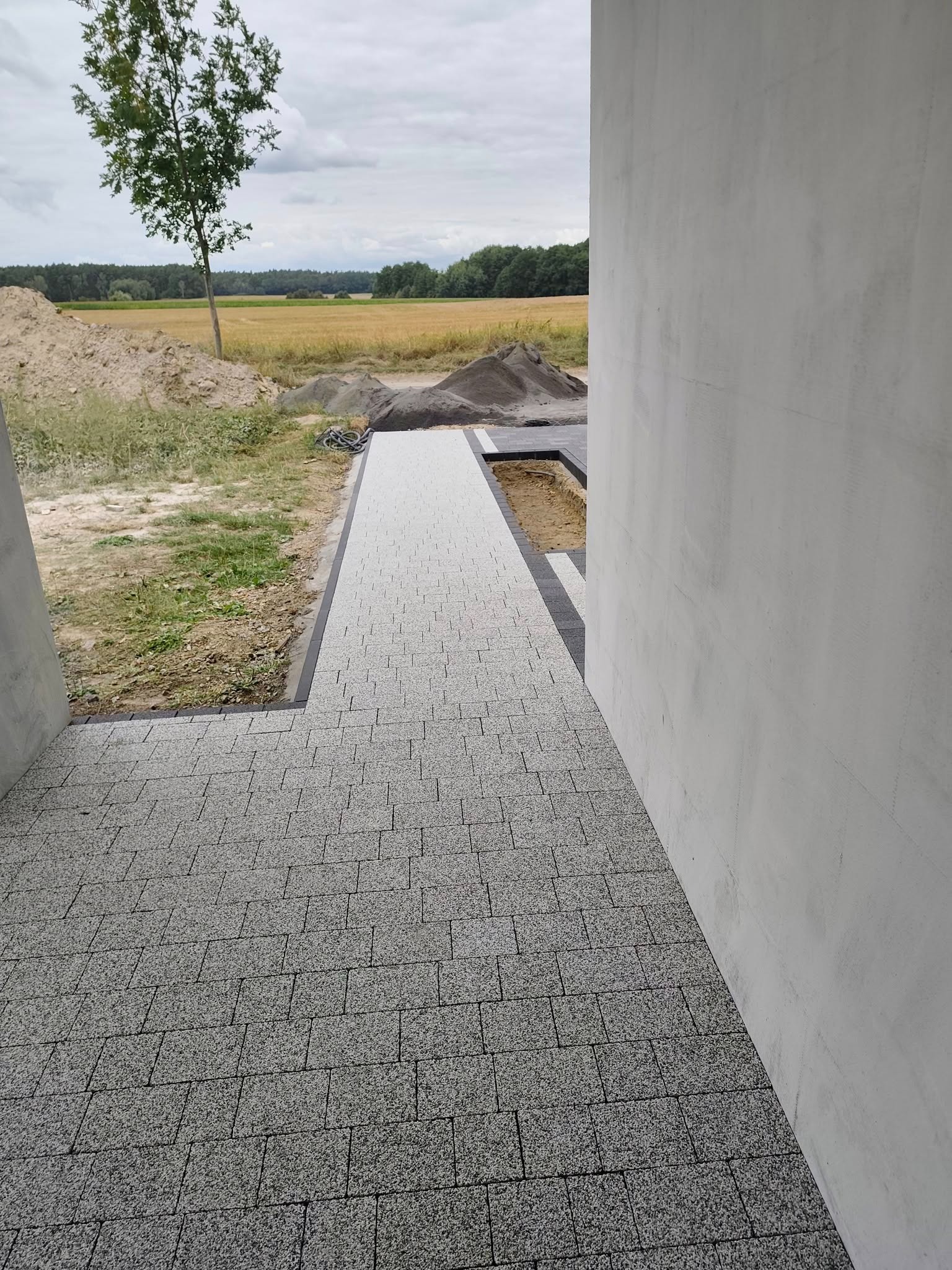 Ułożona kostka brukowa w odcieniach szarości, tworząca ścieżkę z czarnym obramowaniem, prowadząca do budynku. W tle widoczne pole i drzewa.