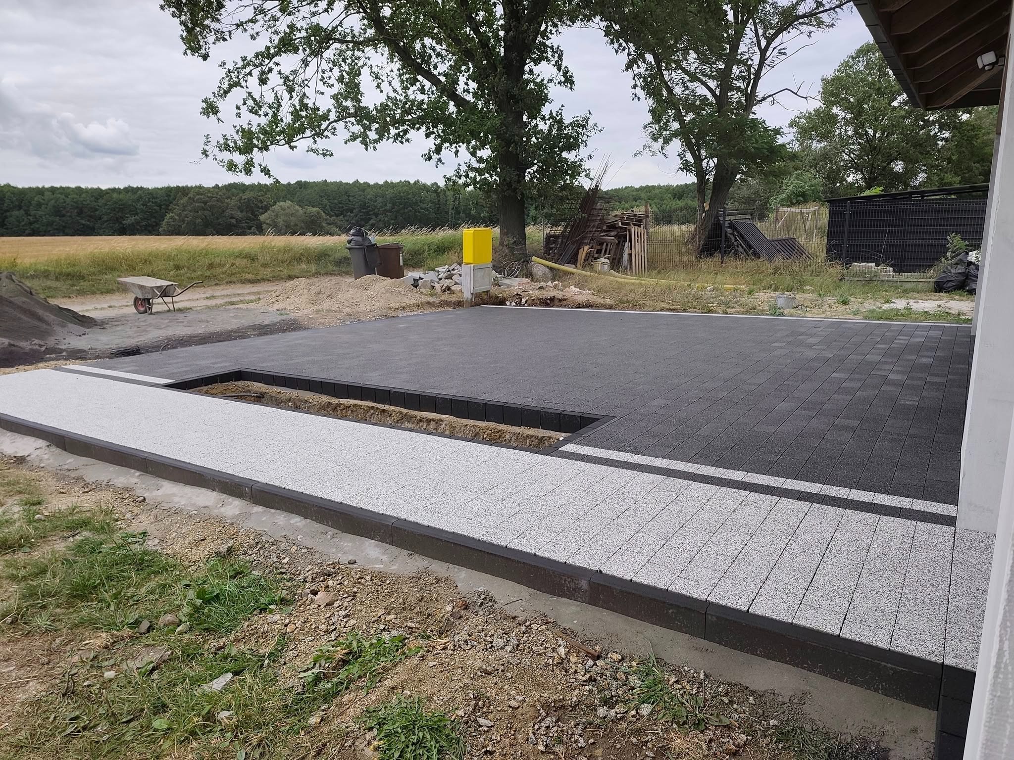 Nowoczesny taras z kostki brukowej w odcieniach szarości, z wyraźnie oddzieloną strefą. W tle widoczne drzewa i polne otoczenie, co podkreśla naturalny charakter przestrzeni.