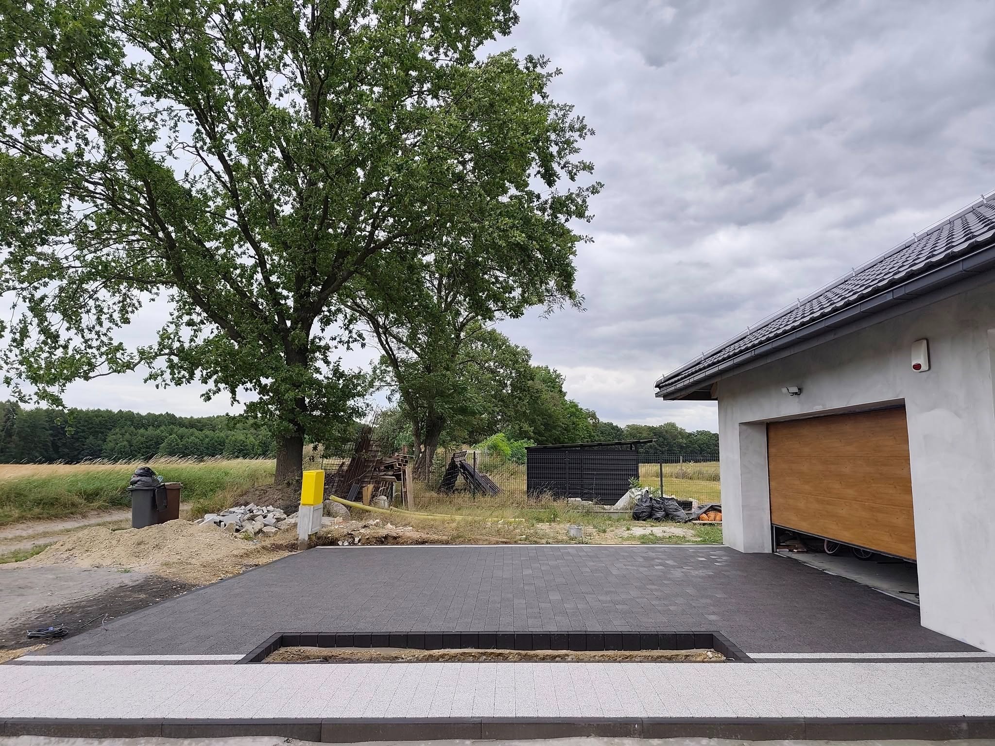 Częściowo ułożony podjazd z kostki w odcieniach szarości, z widocznym obrzeżem w kolorze białym i czarnym, garaż z drewnianą bramą i zielone drzewa w tle.