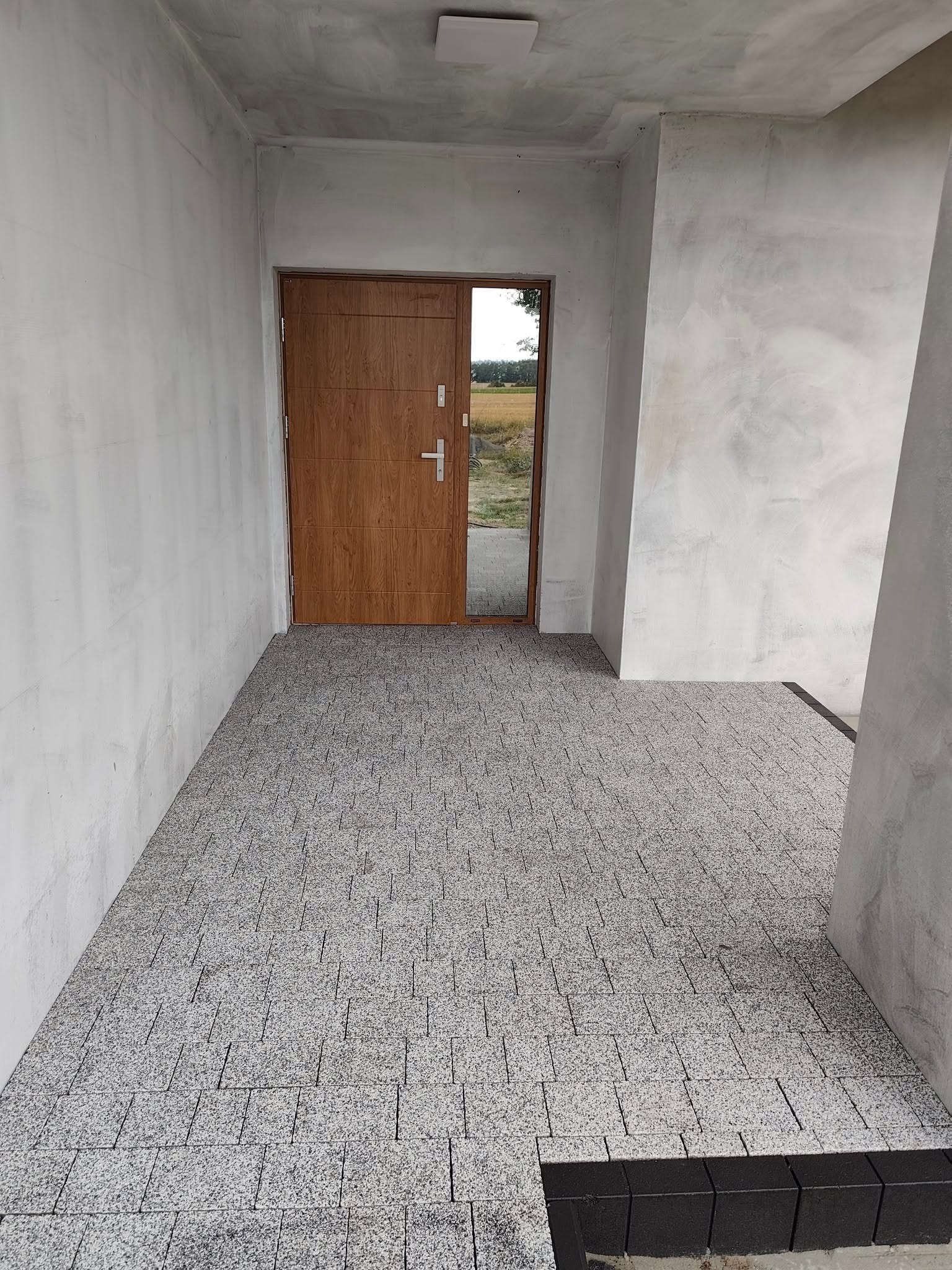 Szara kostka brukowa na zewnątrz budynku, widok na drewniane drzwi wejściowe i surowe ściany. Minimalistyczny styl wykończenia.