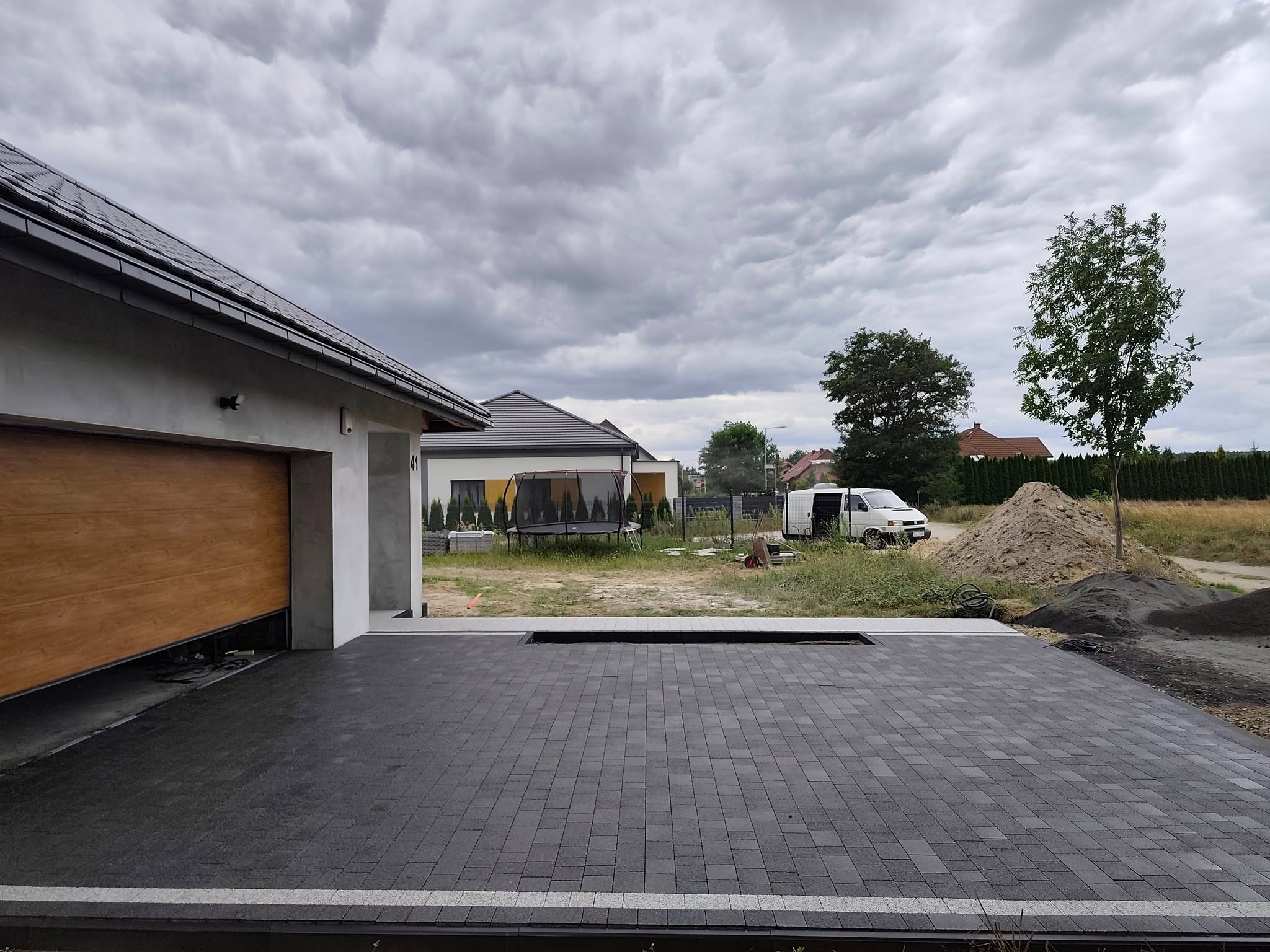 Nowoczesna, ciemnoszara kostka brukowa ułożona na podjeździe przed domem z otwartym garażem. Widoczny fragment ogrodu i zaparkowany biały bus w tle.