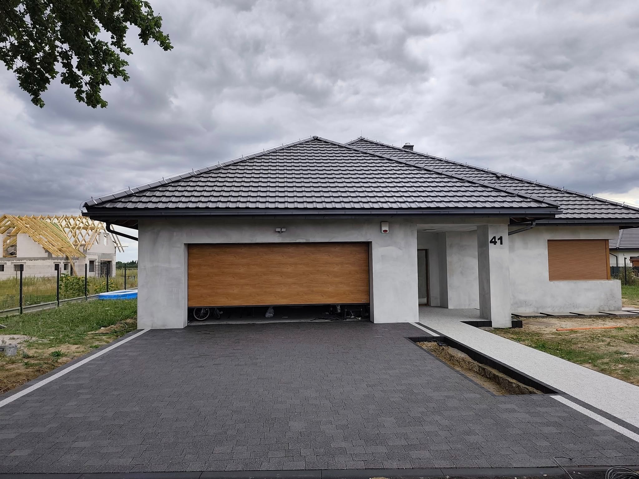 Nowoczesny dom z szarej kostki brukowej i otwartym garażem z drewnianą bramą. Widoczny fragment drenażu. W tle budowa drugiego domu.