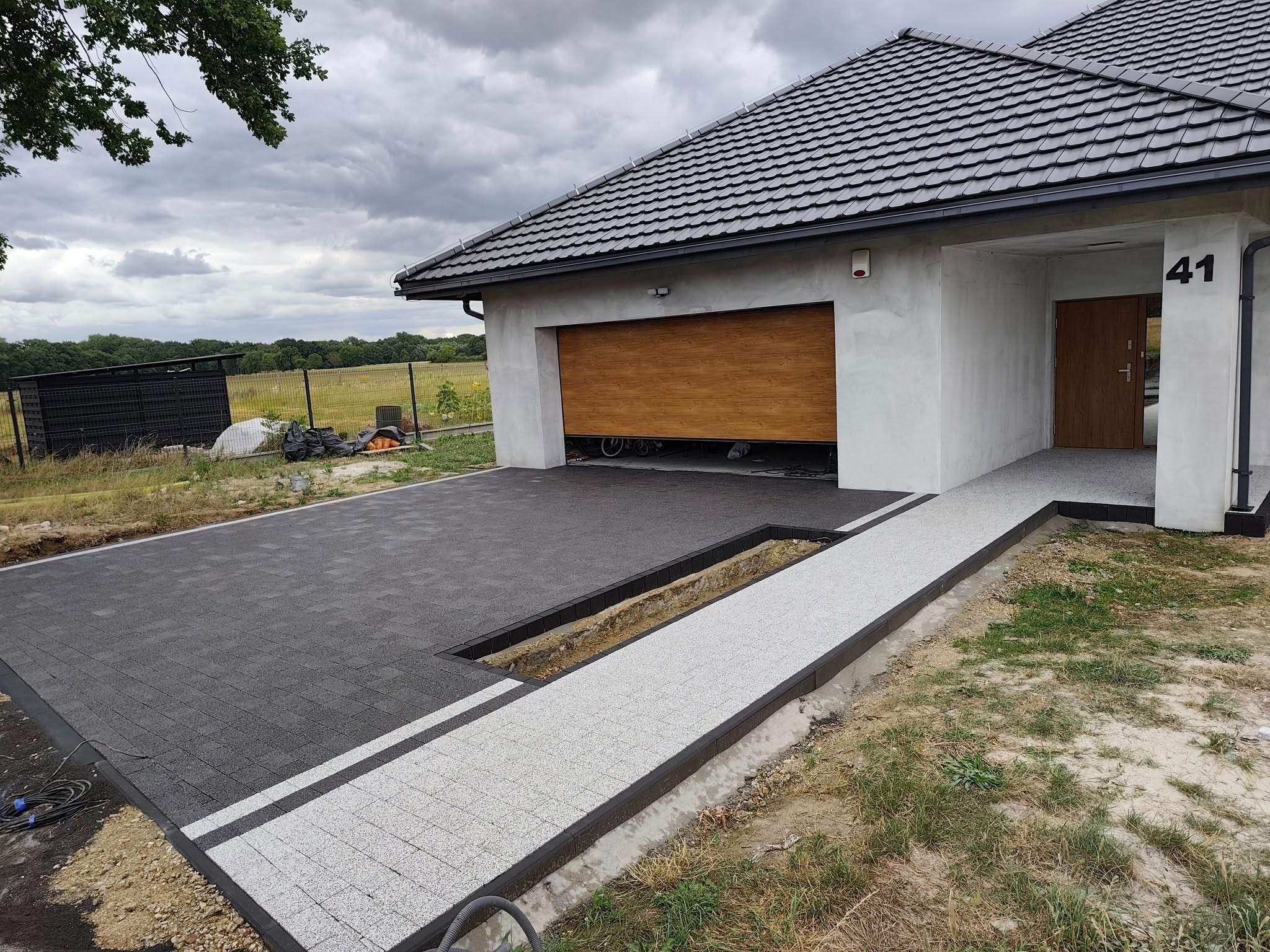 Nowoczesny podjazd z kostki brukowej w odcieniach szarości, z eleganckim obramowaniem i numerem domu 41 w tle. Widoczny fragment garażu z drewnianą bramą.