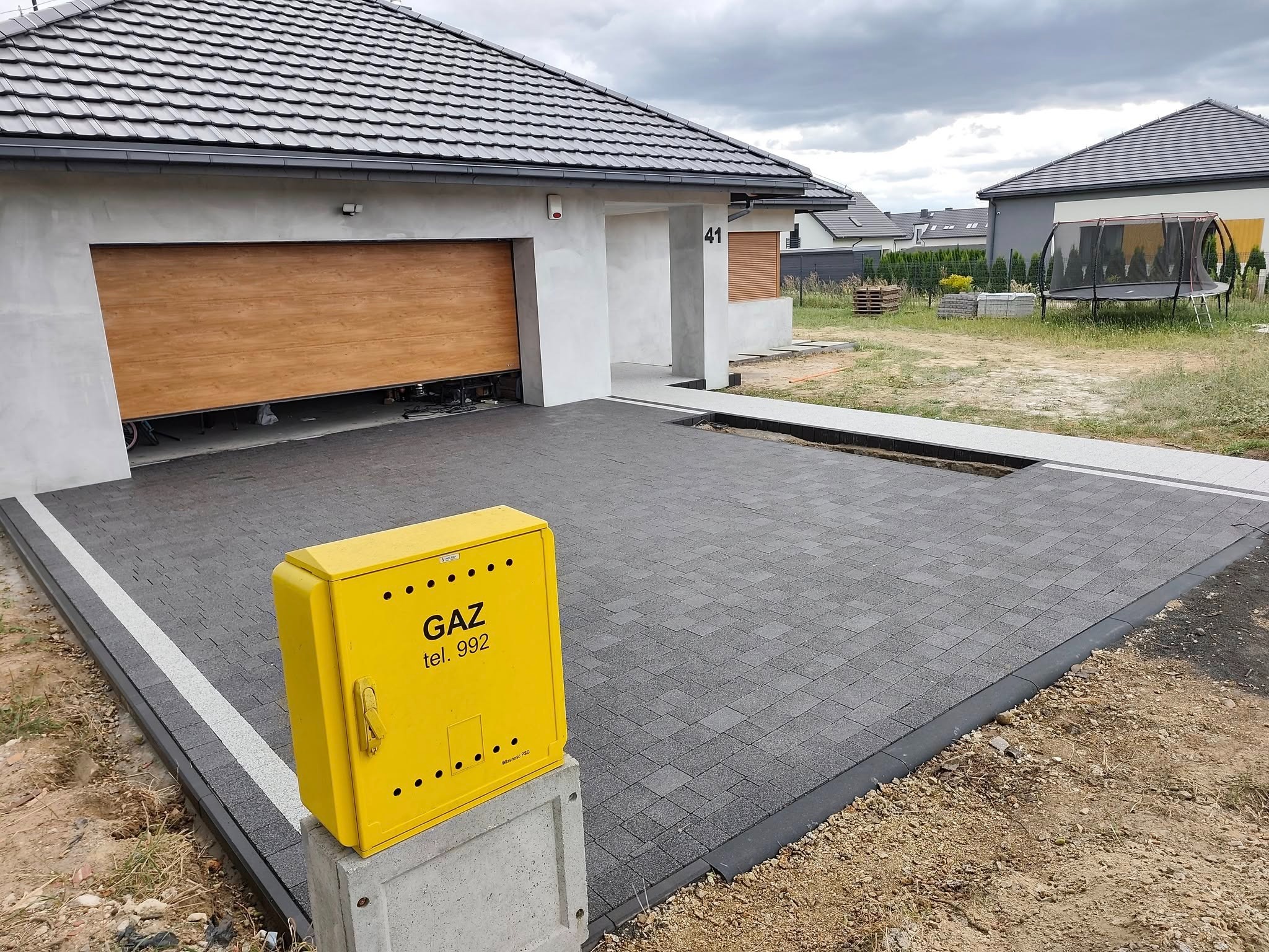 Nowoczesny podjazd z kostki brukowej w odcieniach szarości, z żółtą skrzynką gazową na pierwszym planie i częściowo otwartym garażem w tle.