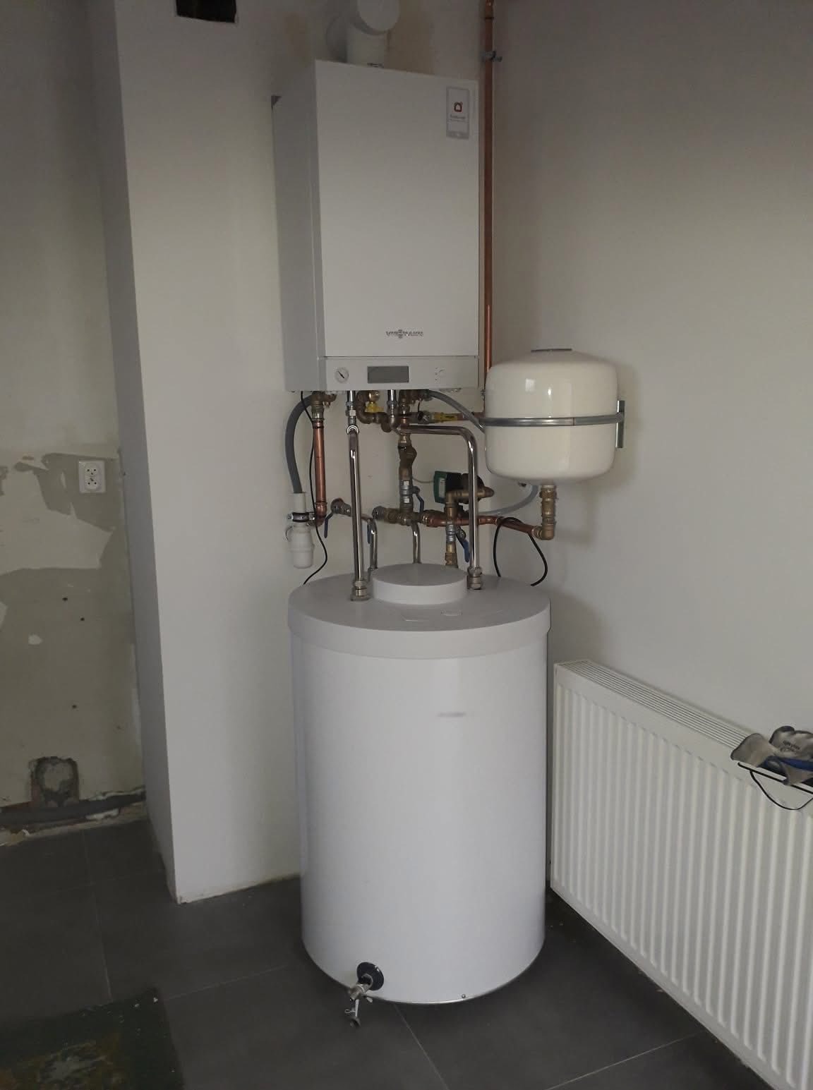 Biały, wiszący piec gazowy Viessmann z podłączonym zasobnikiem ciepłej wody i instalacją hydrauliczną w pomieszczeniu z grzejnikiem.