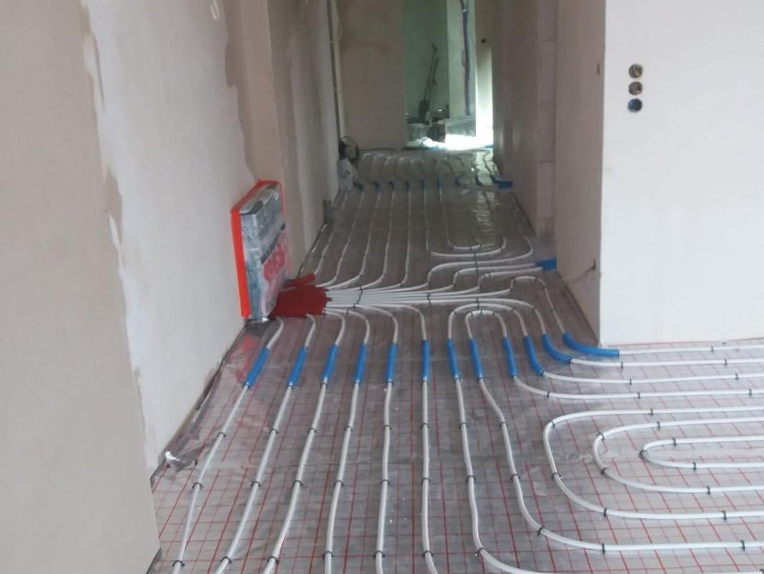 Białe rury ogrzewania podłogowego z niebieskimi łącznikami, ułożone na siatce w surowym wnętrzu z widocznymi śladami prac wykończeniowych. Widok z perspektywy.