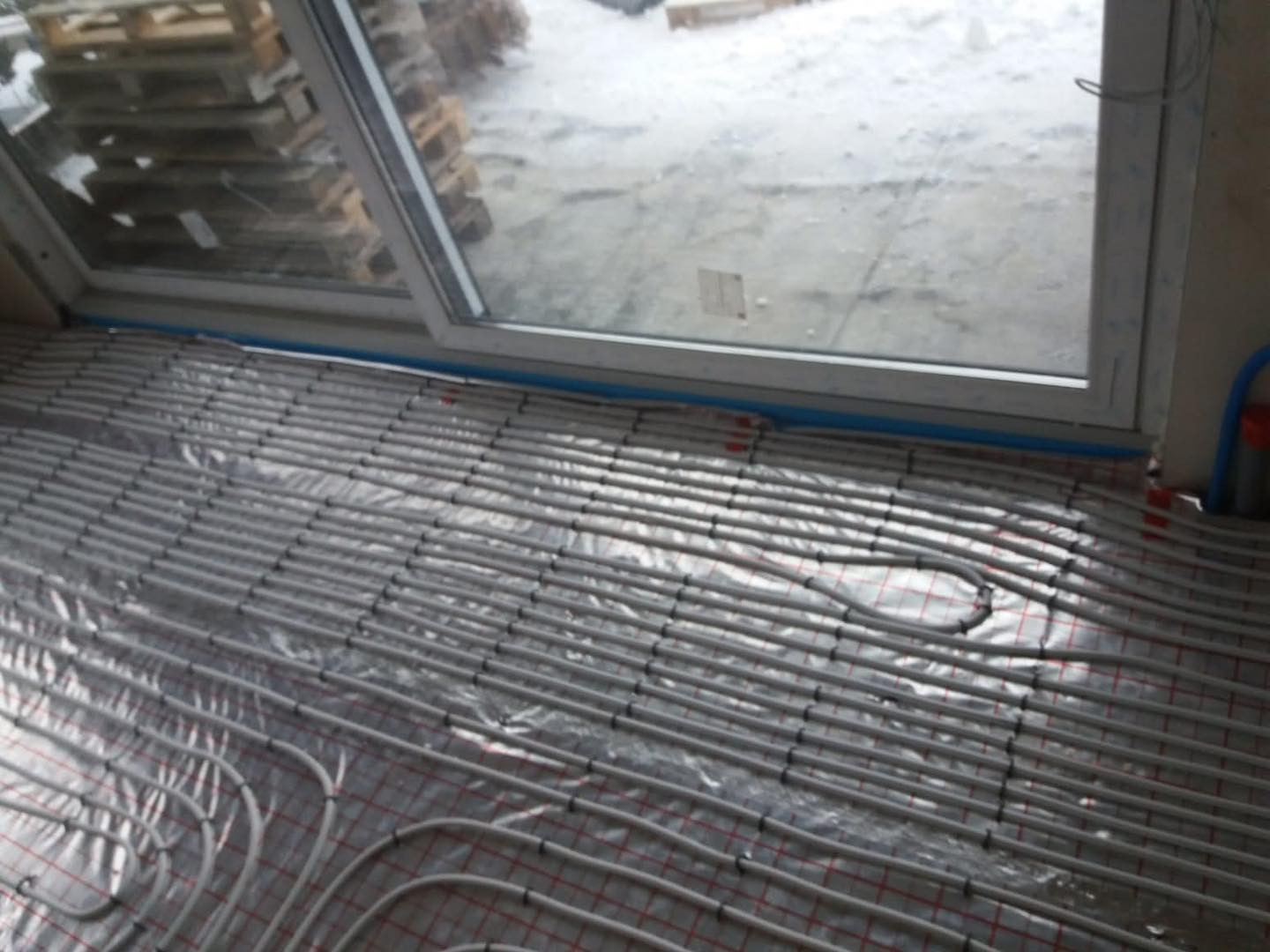 Ułożone rury ogrzewania podłogowego na folii z siatką, widok z góry, przy oknie. Instalacja przed zalaniem posadzki, etap prac.