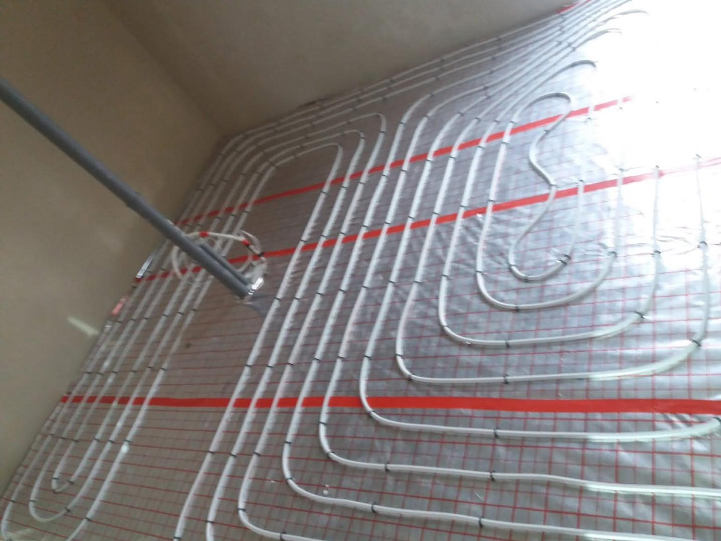 Ułożone białe rury ogrzewania podłogowego na folii z czerwoną siatką, z szarą rurą doprowadzającą po lewej. Widok pod kątem, surowy stan.