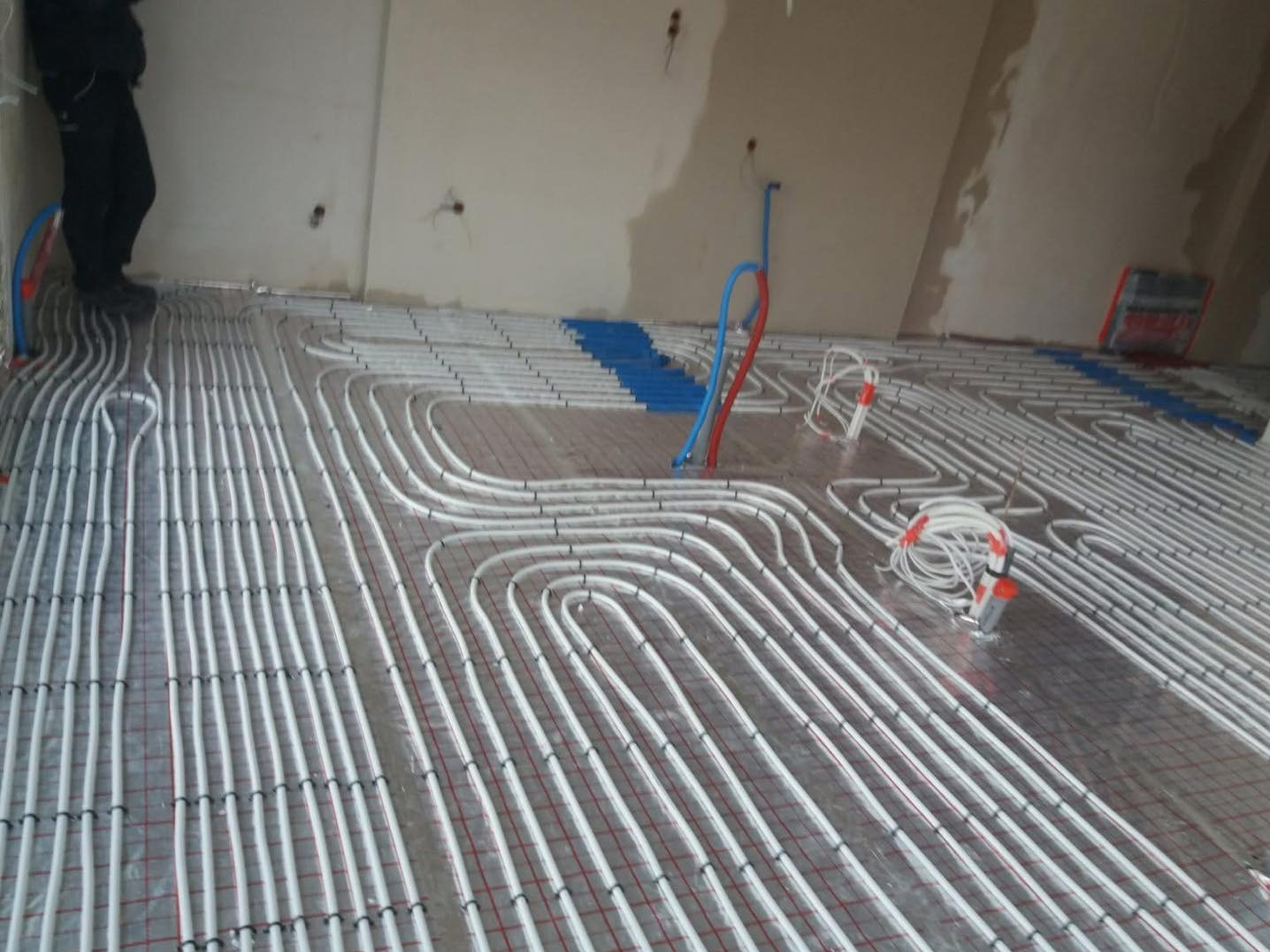 Białe rury ogrzewania podłogowego ułożone na siatce, widoczne niebieskie i czerwone rury przy ścianie. W tle osoba w czarnych spodniach.