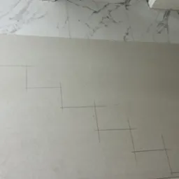 Płyta gipsowo-kartonowa z naniesionym ołówkiem schematem schodkowym, leżąca na marmurowej posadzce. Widoczne fragmenty drabiny malarskiej.