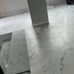 Białe płytki podłogowe z marmurkowym wzorem, fragment schodów z szarego kamienia, szara kolumna w pomieszczeniu z oknem i kartonem w tle.