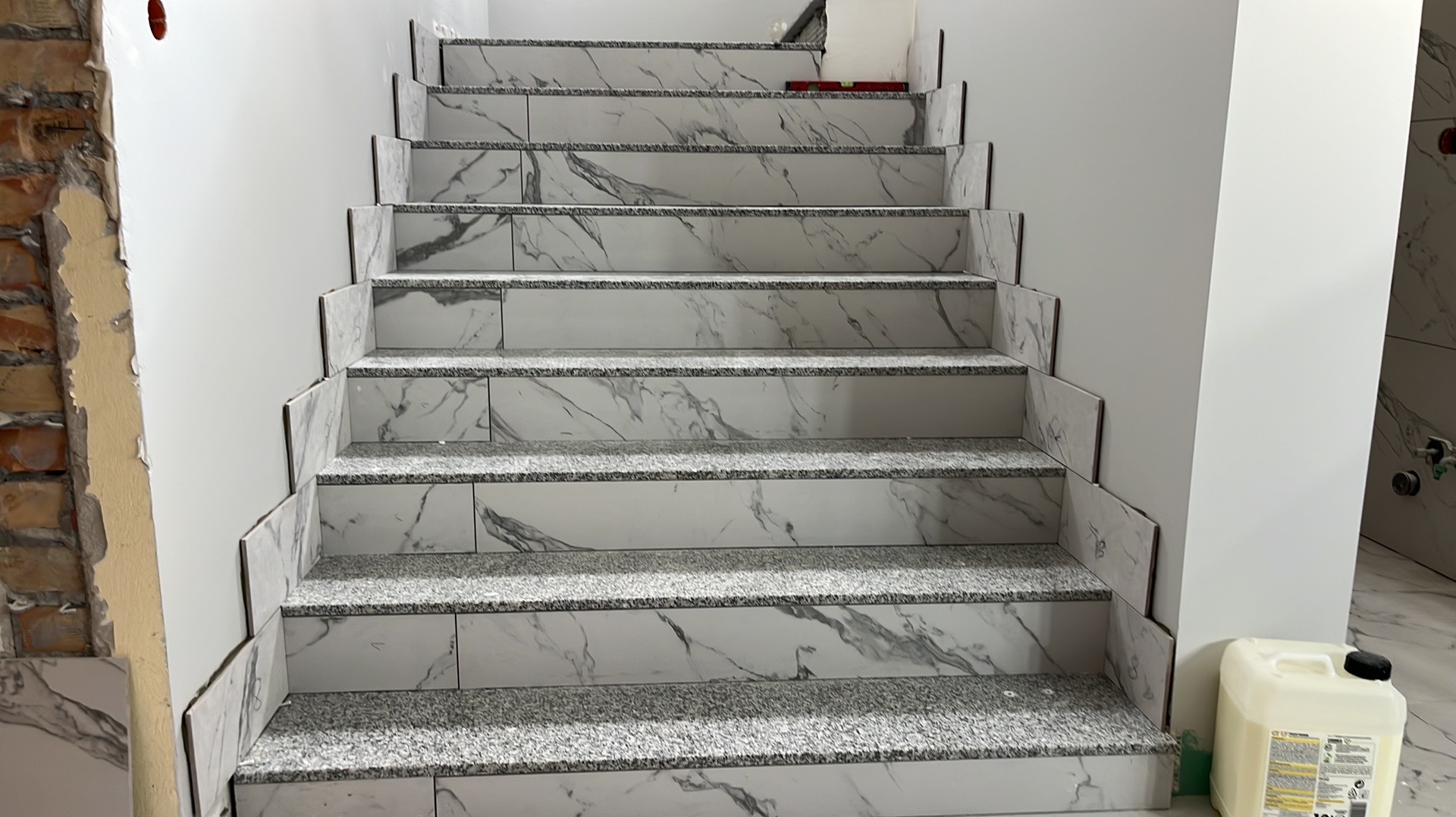 Wykończone schody z białymi płytkami imitującymi marmur i szarymi stopnicami. Widoczne niedokończone fragmenty ściany i pojemnik z klejem.