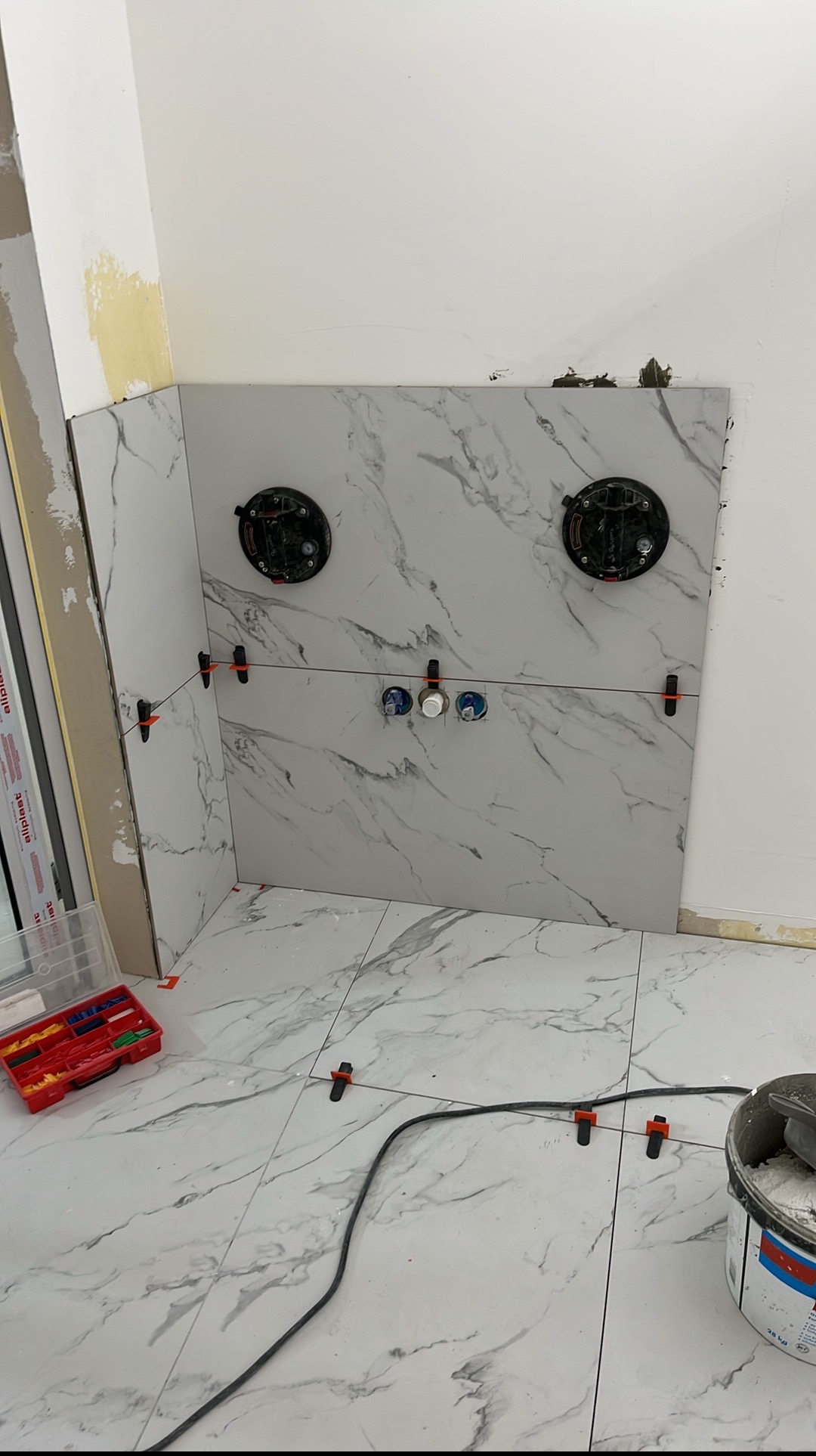 Montaż marmurowych płytek w łazience z użyciem przyssawek i klipsów poziomujących. Widoczne narzędzia i puszki instalacyjne. Etap prac wykończeniowych.