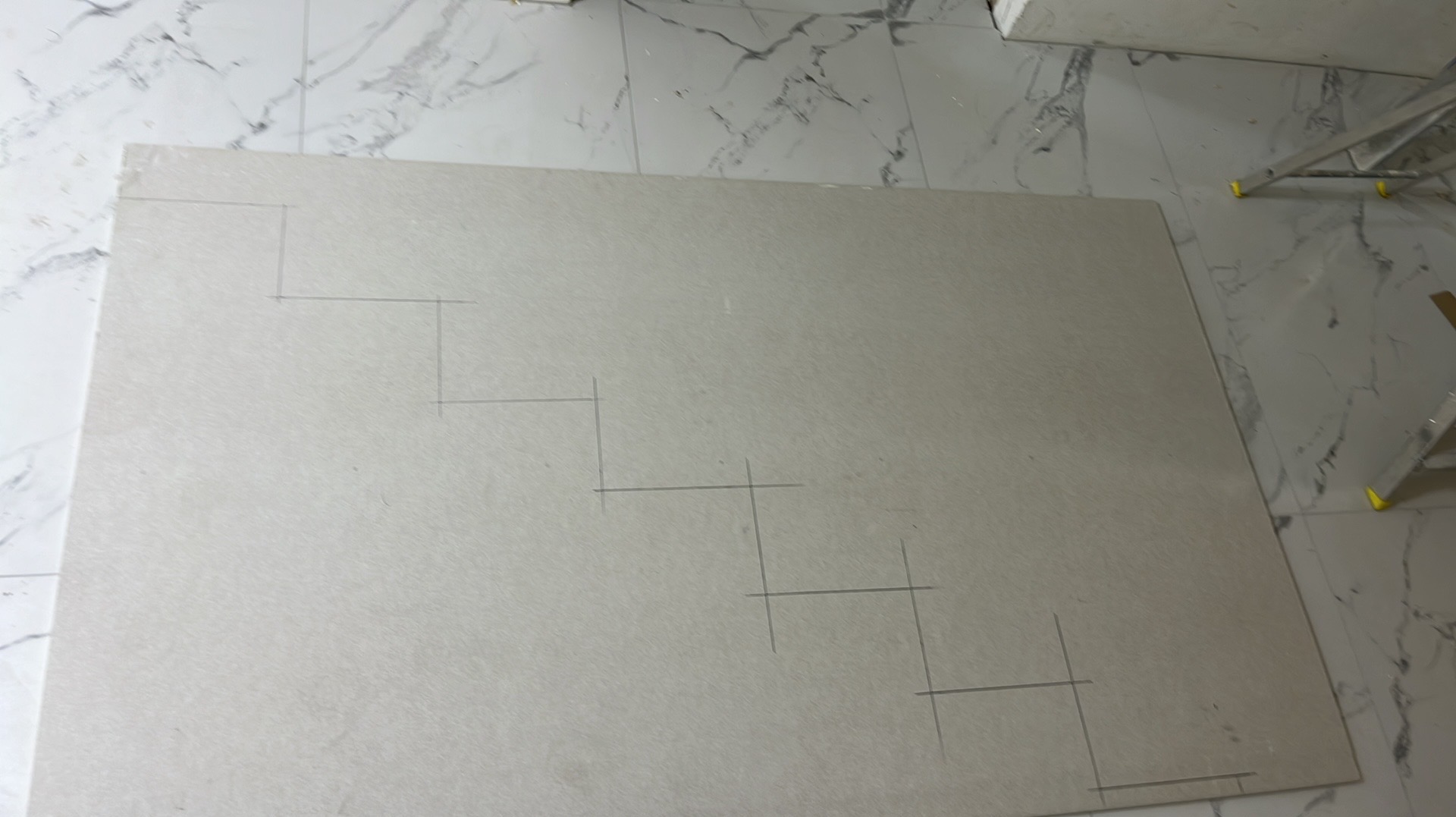 Płyta gipsowo-kartonowa z naniesionym ołówkiem schematem schodkowym, leżąca na marmurowej posadzce. Widoczne fragmenty drabiny malarskiej.