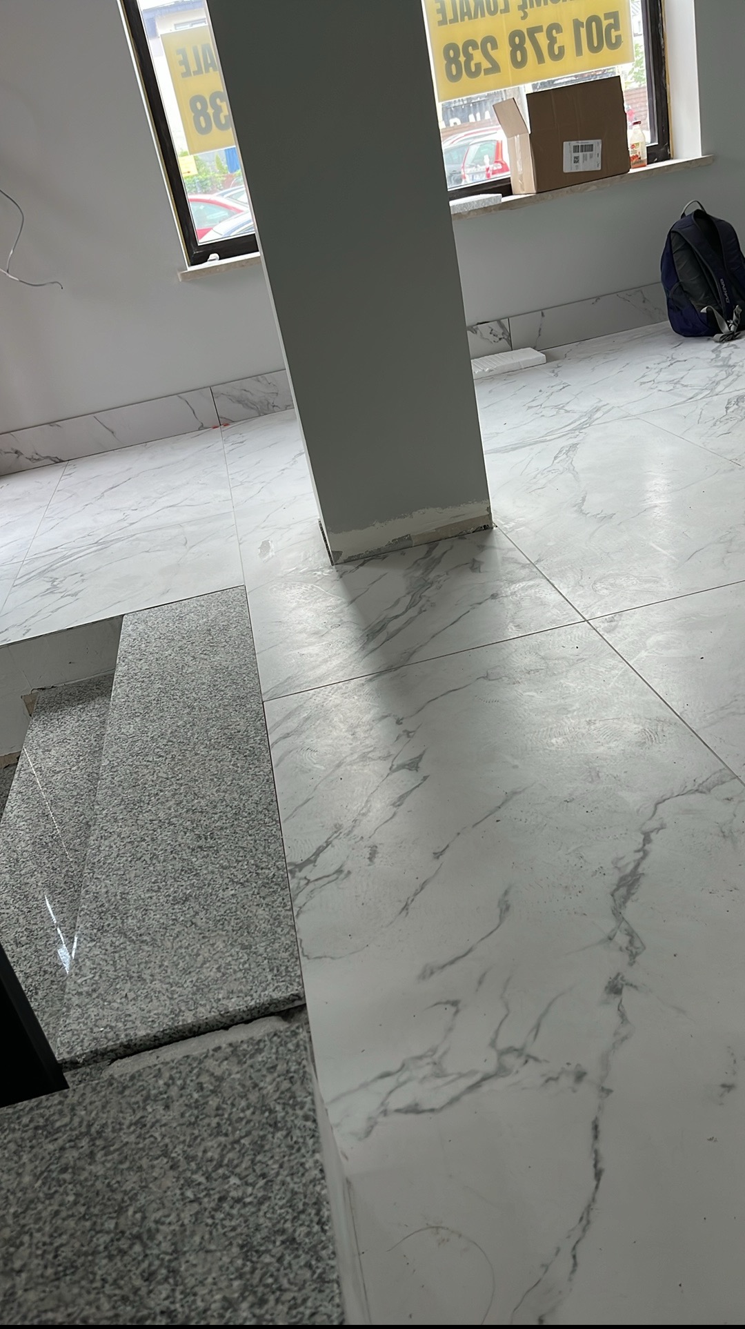 Białe płytki podłogowe z marmurkowym wzorem, fragment schodów z szarego kamienia, szara kolumna w pomieszczeniu z oknem i kartonem w tle.