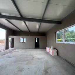 TynkMaksBud - Wnętrze w stanie surowym: szare tynki, betonowa podłoga, białe okna, metalowa konstrukcja dachu. Materiały budowlane w rogu pomieszczenia.