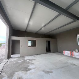 TynkMaksBud - Wnętrze w stanie surowym: ściany po tynkowaniu, widoczne okno, sufit z metalową konstrukcją, podłoga betonowa. Na ścianie leżą zapakowane materiały budowlane.