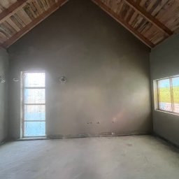 TynkMaksBud - Surowe wnętrze po tynkowaniu, z drewnianym sufitem w kształcie litery A, widoczne okna i szare ściany. Wnętrze w trakcie prac wykończeniowych.