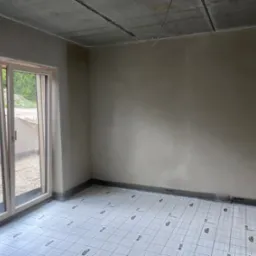 Pokój po tynkowaniu ścian, z oknem balkonowym i przygotowaną podłogą z izolacją, gotowy do dalszych prac wykończeniowych. Widoczne surowe wykończenie sufitu.
