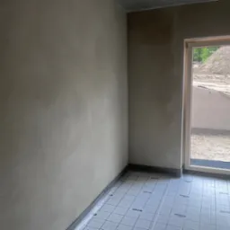 Ściana po tynkowaniu z widoczną siatką pod ogrzewanie podłogowe i oknem z widokiem na zewnątrz. Surowy betonowy sufit i widoczne elementy wykończeniowe.