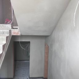 TynkMaksBud - Wnętrze w trakcie prac: świeżo otynkowane ściany i sufit, widoczne schody w surowym stanie oraz instalacja elektryczna w otworze drzwiowym. Podłoga zabezpieczona.