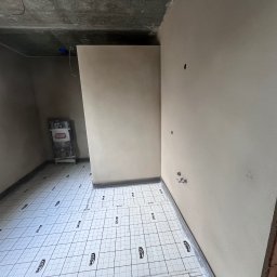 TynkMaksBud - Wnętrze w trakcie prac wykończeniowych: ściany pokryte jasną gładzią, podłoga zabezpieczona folią ochronną z nadrukiem, widoczny narożnik z cegły.