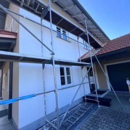 TynkMaksBud - Dom jednorodzinny w rusztowaniach po świeżym, białym tynkowaniu. Widoczne okna, dachówka i fragment chodnika z kostki. Niebieskie niebo w tle.