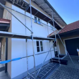 Dom jednorodzinny w rusztowaniach po świeżym, białym tynkowaniu. Widoczne okna, dachówka i fragment chodnika z kostki. Niebieskie niebo w tle.